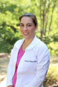 The image shows a woman wearing a white lab coat standing outdoors with trees behind her.