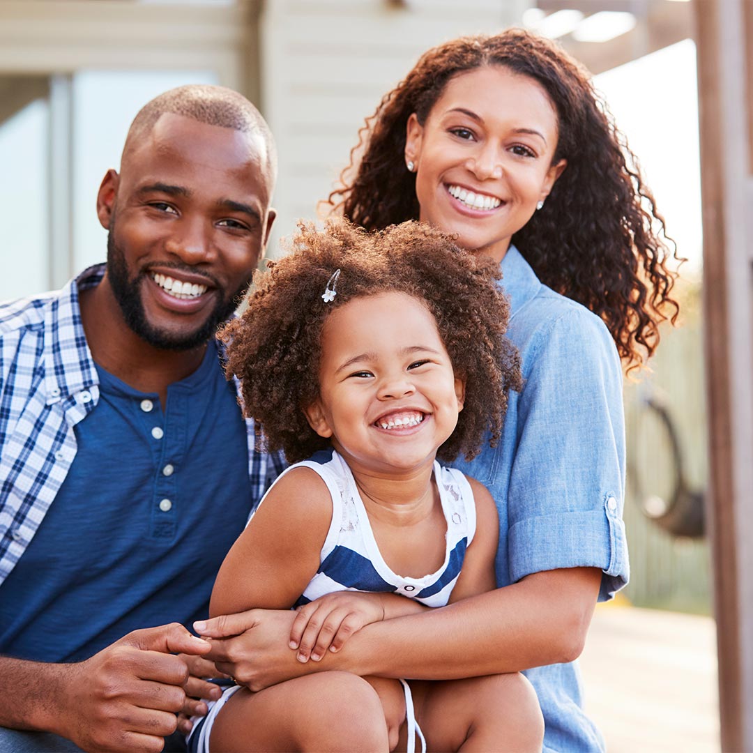 In this image, there are four individuals  an adult male, an adult female, a young child, and a baby. They appear to be a family enjoying a sunny day outdoors, with the adults smiling at the camera and the children looking away. The setting suggests a casual, relaxed atmosphere, possibly during a family outing or vacation.