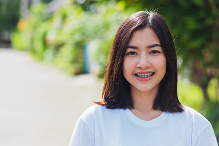 A young woman with short hair smiling at the camera, wearing a white t-shirt, standing outdoors in front of a building with greenery.