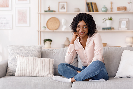 A young woman with curly hair is sitting on a couch indoors, smiling at the camera.